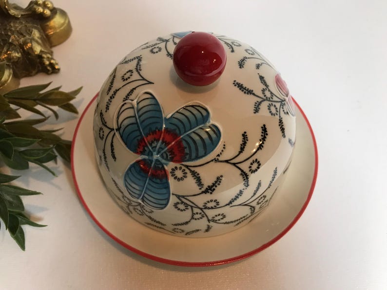 May include: A white ceramic butter dish with a domed lid and a matching plate. The lid features a large blue and red floral design, a red knob, and black vine-like accents. The plate has a red rim.