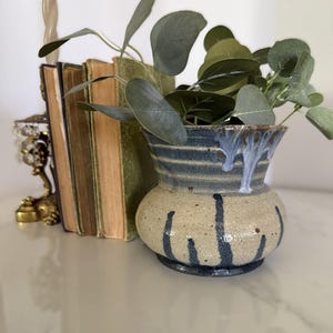 May include: A ceramic vase with a rounded base and flared rim, featuring a speckled beige glaze with dark blue vertical drip patterns and horizontal bands near the top, containing a sprig of green eucalyptus leaves with rounded foliage.