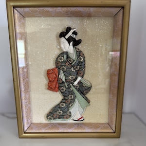 May include: A framed three-dimensional fabric wall art piece featuring a figure in a dark patterned kimono with floral motifs, a bright orange obi, and a pale green underlayer. The figure has dark hair and is set against a textured cream background.