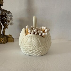 May include: Cream-colored swan-shaped match holder filled with matches. The ceramic holder has a textured feather design and a curved neck. The matches have white tips. A decorative brass candle holder with crystal accents is partially visible in the background.