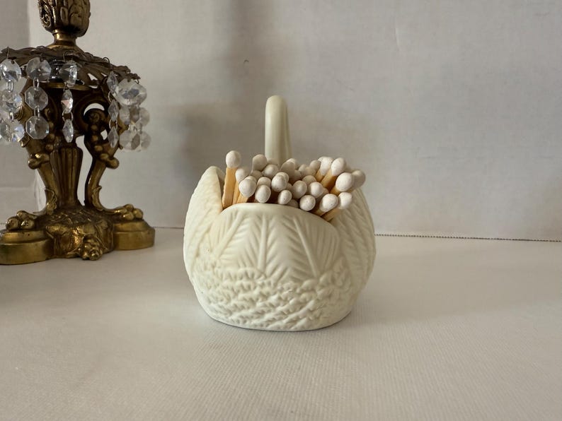 May include: Cream-colored swan-shaped match holder filled with matches. The ceramic holder has a textured feather design and a curved neck. The matches have white tips. A decorative brass candle holder with crystal accents is partially visible in the background.