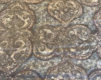 May include: Close-up of a fabric with a repeating pattern of ornate, scroll-like designs in shades of brown, beige, and blue-gray. The pattern has a vintage or antique aesthetic, with a textured appearance.