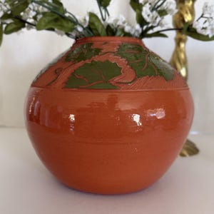 Peut inclure: Un vase en céramique de couleur terre cuite avec un corps rond et une large ouverture. Le vase présente un motif de feuilles vertes autour du bord. Des fleurs blanches et du feuillage vert sont disposés à l'intérieur du vase. Un chandelier doré est visible en arrière-plan.