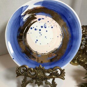 May include: A white ceramic plate with a blue and gold abstract design. The plate has a pink and white center with gold brushstrokes and blue dots. The plate is on a gold metal stand.