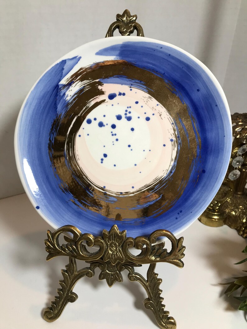 May include: A white ceramic plate with a blue and gold abstract design. The plate has a pink and white center with gold brushstrokes and blue dots. The plate is on a gold metal stand.