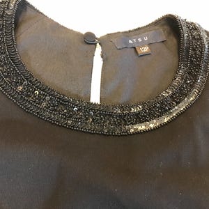 May include: Close-up of a black dress neckline. The neckline is embellished with a beaded design. The dress has a small keyhole opening in the front. The label reads "ATSU 12P".