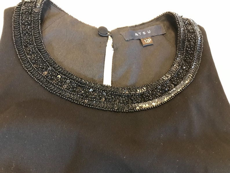 May include: Close-up of a black dress neckline. The neckline is embellished with a beaded design. The dress has a small keyhole opening in the front. The label reads "ATSU 12P".