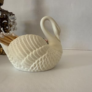 May include: A cream-colored swan-shaped match holder. The ceramic holder features detailed feather textures and holds a bundle of wooden matches. The swan's neck curves gracefully upwards. A decorative brass object with crystals is in the background.