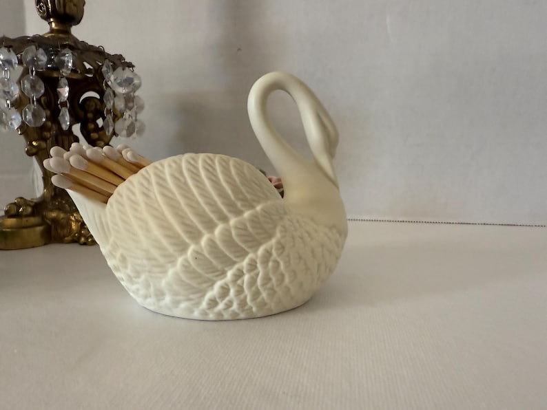 May include: A cream-colored swan-shaped match holder. The ceramic holder features detailed feather textures and holds a bundle of wooden matches. The swan's neck curves gracefully upwards. A decorative brass object with crystals is in the background.