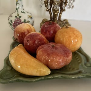May include: A selection of stone fruit sculptures, including apples, a peach, and a banana, presented on a green ceramic tray. The fruit sculptures are in shades of red, orange, and yellow. A decorative vase and a crystal chandelier are in the background.