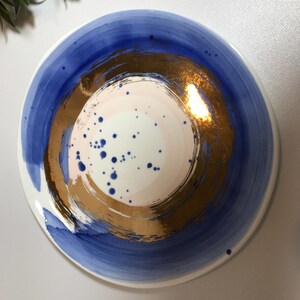 May include: A white ceramic plate with a blue and gold design. The plate has a large, circular, gold design in the center with a pink and white center. The gold design is surrounded by a blue ring.