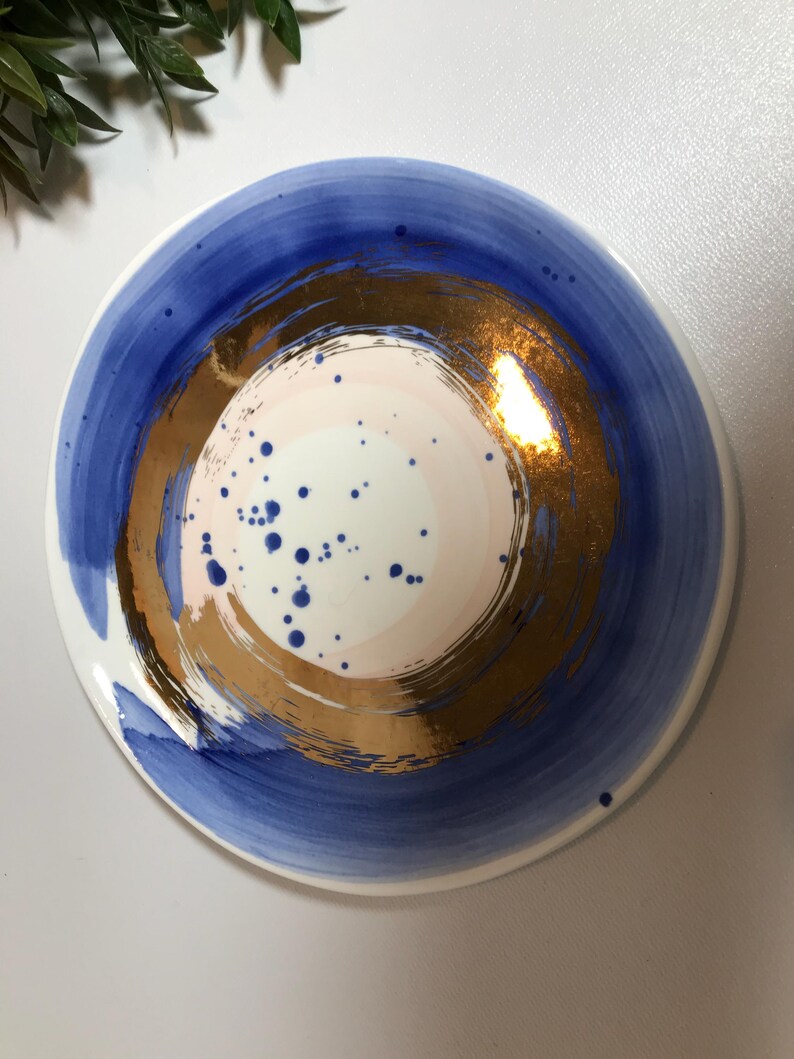 May include: A white ceramic plate with a blue and gold design. The plate has a large, circular, gold design in the center with a pink and white center. The gold design is surrounded by a blue ring.