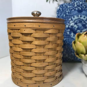 May include: A cylindrical, light brown woven basket with a wooden lid and a small, metallic knob. The basket is made of interwoven strips of wood, creating a textured surface. The lid is circular and fits snugly on top. A decorative storage container.