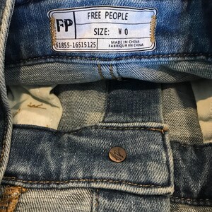 Peut inclure: Une &eacute;tiquette en jean bleu avec le texte "FREE PEOPLE" et "SIZE: W 0" en blanc. L'&eacute;tiquette comprend &eacute;galement le texte "MADE IN CHINA" et "FABRIQUER EN CHINE" en noir. L'&eacute;tiquette a des coutures brunes autour des bords et le texte "FP" en blanc sur un fond brun.