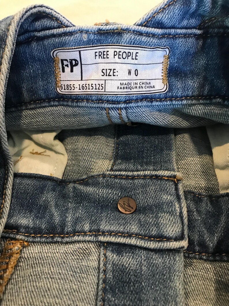 Peut inclure: Une &eacute;tiquette en jean bleu avec le texte "FREE PEOPLE" et "SIZE: W 0" en blanc. L'&eacute;tiquette comprend &eacute;galement le texte "MADE IN CHINA" et "FABRIQUER EN CHINE" en noir. L'&eacute;tiquette a des coutures brunes autour des bords et le texte "FP" en blanc sur un fond brun.