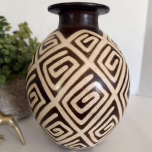May include: A decorative ceramic vase with a brown rim and a cream-colored body. The body is adorned with a repeating geometric pattern in dark brown. The vase has a rounded shape and a narrow neck.