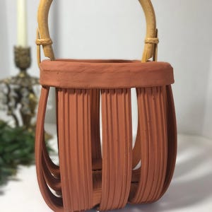 May include: A terracotta-colored clay basket featuring a series of vertical, ribbed slats that form a cage-like structure. The basket is topped with a thick, solid rim and includes a curved, light-colored bamboo handle attached with woven accents.