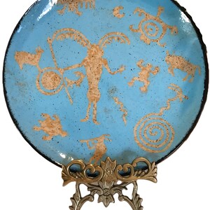 May include: A round, light blue decorative plate with tan-colored petroglyph-style figures and symbols. The plate has a black rim and is displayed on a decorative brass stand. The figures include animals and human-like forms.