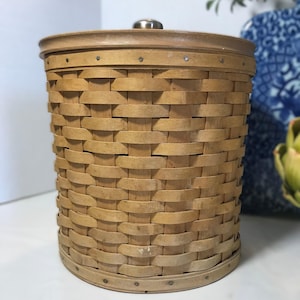 May include: A cylindrical, light brown woven basket with a matching lid. The basket is made of interwoven wooden strips, with a metal knob on the lid. The rim and base are reinforced with a darker wood trim and small metal rivets.