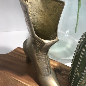 May include: A gold metal boot shaped vase with a high heel. The boot is decorated with a floral design and has a small opening at the top for flowers.