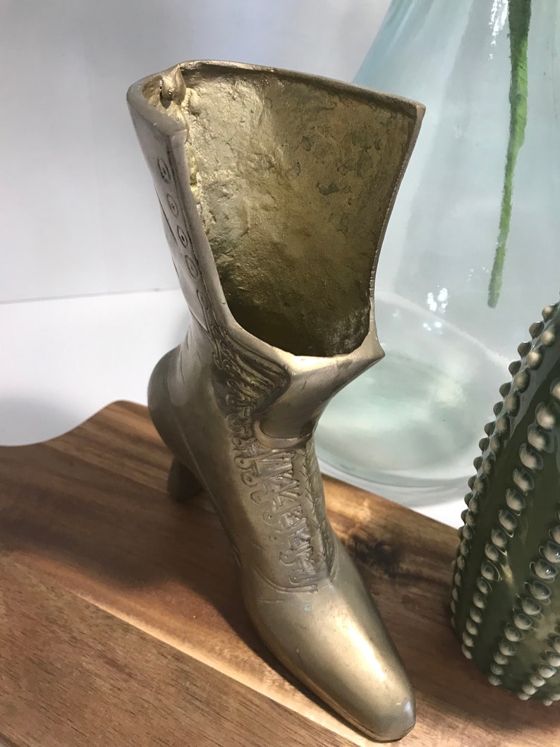 May include: A gold metal boot shaped vase with a high heel. The boot is decorated with a floral design and has a small opening at the top for flowers.