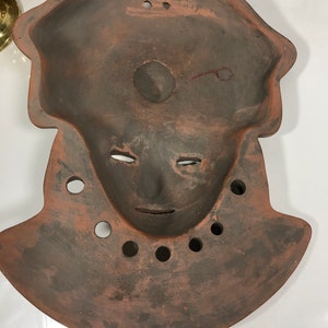May include: A terracotta mask with a human face and a rounded, slightly pointed top. The mask has a hole in the center of the face and several holes around the neck.