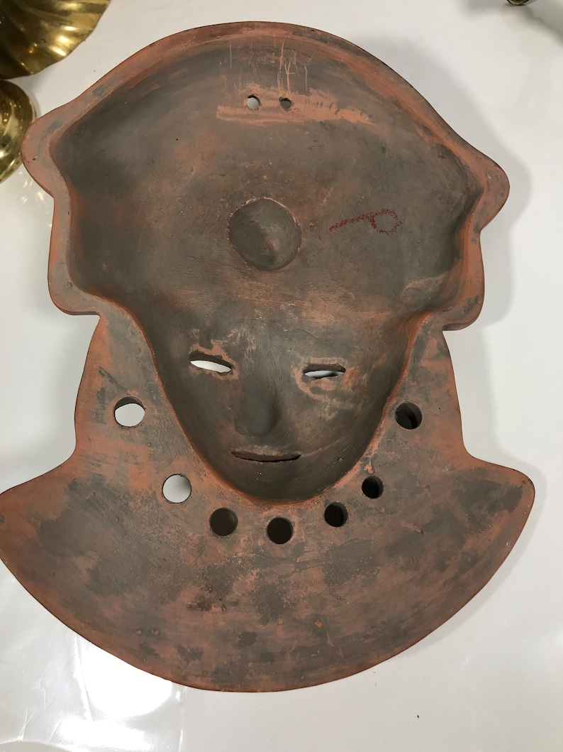 May include: A terracotta mask with a human face and a rounded, slightly pointed top. The mask has a hole in the center of the face and several holes around the neck.