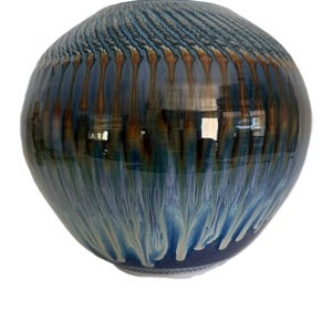 May include: A round ceramic vase with a glossy finish. The vase features a blend of blue, brown, and cream colors. The upper portion has a textured pattern, while the lower part has a flowing, dripped design.