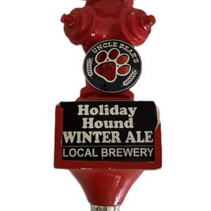May include: A red beer tap shaped like a fire hydrant, with the words "Holiday Hound Winter Ale" and "Local Brewery" on a black label. The tap has a silver base and a circular emblem with a paw print and the words "Uncle Bear's".