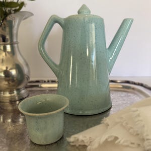 May include: A light blue ceramic teapot with a speckled finish and a matching small cup. The teapot features a tall, cylindrical body, a pointed lid, a curved handle, and a straight spout. The set is presented on a reflective silver tray.