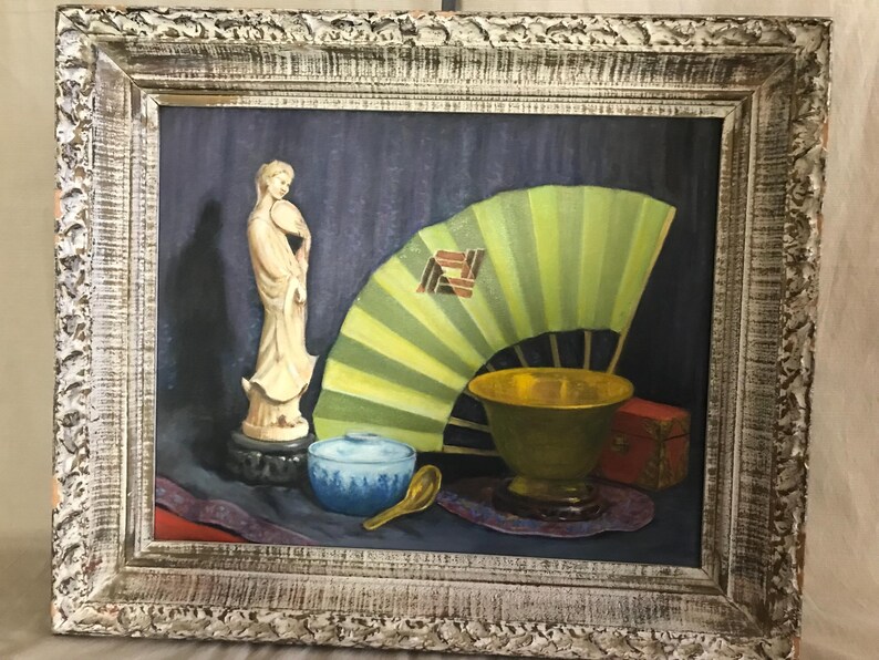 May include: A still life painting of a white porcelain figurine, a green and yellow fan, a blue and white bowl, a gold bowl, and a red box. The painting is framed in a white wooden frame.