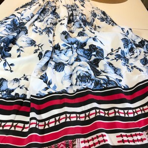 May include: A black top and a long white skirt with a blue floral pattern. The skirt has a red, black, and white striped border and a straw hat. The outfit is laid out on a white surface.