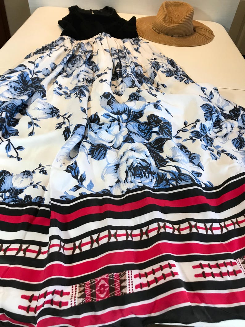 May include: A black top and a long white skirt with a blue floral pattern. The skirt has a red, black, and white striped border and a straw hat. The outfit is laid out on a white surface.
