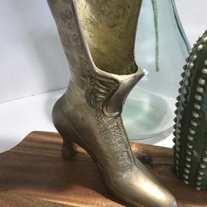 May include: A gold-colored metal boot-shaped vase with a high heel. The boot has a lace-up design and is sitting on a wooden surface.