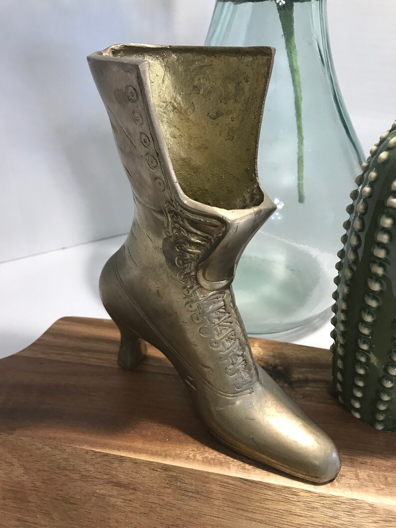 May include: A gold-colored metal boot-shaped vase with a high heel. The boot has a lace-up design and is sitting on a wooden surface.