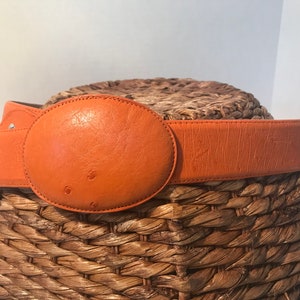 Op de afbeelding: Een feloranje leren riem met een grote ovale gesp. De riem ligt op een geweven strooppervlak.