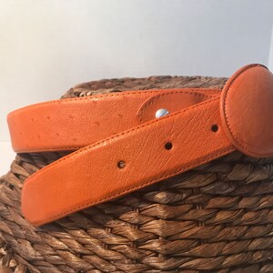 Op de afbeelding: Een feloranje leren riem met een ronde gesp. De riem is gemaakt van struisvogelhuid en heeft een subtiel patroon.