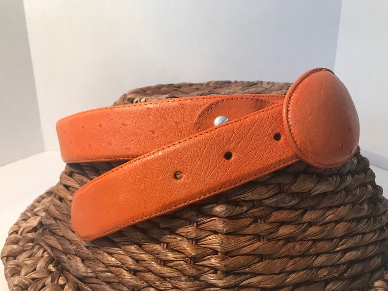 Op de afbeelding: Een feloranje leren riem met een ronde gesp. De riem is gemaakt van struisvogelhuid en heeft een subtiel patroon.