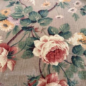 May include: Close-up of a floral fabric featuring large, detailed roses in shades of red and cream, surrounded by green leaves and smaller flowers in yellow and white. The background is a neutral beige.