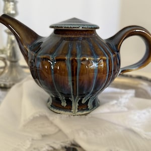May include: A ceramic teapot featuring a glossy brown glaze with vertical blue drip patterns. The teapot has a fluted body, a curved handle, a pointed spout, and a matching lid with a conical top. The surface has a reflective, multi-toned finish.