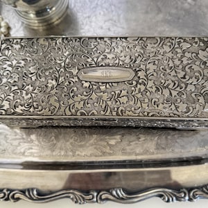 Puede incluir: Una caja rectangular de metal plateado con un intrincado patrón floral y de volutas en relieve en la tapa y los laterales. El centro de la tapa muestra una placa ovalada lisa y alargada grabada con las letras del monograma KJB.