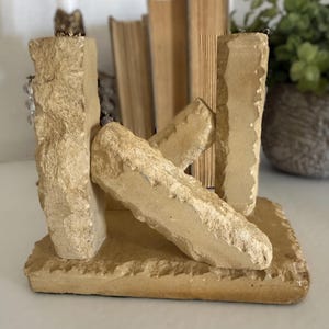 May include: A tan-colored stone bookend featuring a rectangular base with two vertical pillars and a diagonal support beam connecting them. The surface of the stone has a rough, textured, and chiseled appearance, showcasing natural variations in the light beige color.