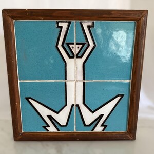 May include: A square wall art piece featuring four glossy turquoise ceramic tiles set within a dark brown wooden frame. The tiles display a stylized white figure with black outlines, depicted with arms raised and legs bent in a geometric design.