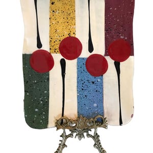 May include: A rectangular ceramic decorative plate with a colourful design. The plate features vertical stripes in white, yellow, blue, green, and burgundy, with red circular accents and black stems. It is displayed on a decorative metal stand.