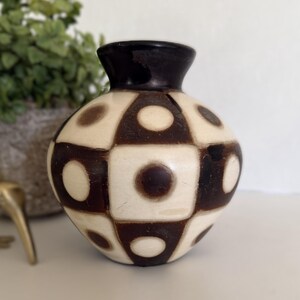 May include: A decorative ceramic vase with a rounded body and a dark brown rim. The vase features a geometric pattern of alternating cream-colored squares and dark brown squares, each with a central brown dot. The vase is a unique home decor item.