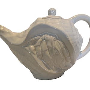 May include: A white ceramic teapot with a seashell design. The teapot features a detailed relief of a hermit crab inside the shell, with a curved handle and a spout. The lid has a small knob.