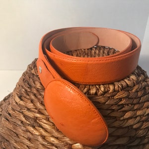 Op de afbeelding: Een feloranje leren riem met een grote, ronde gesp. De riem is opgerold en ligt op een geweven mand.