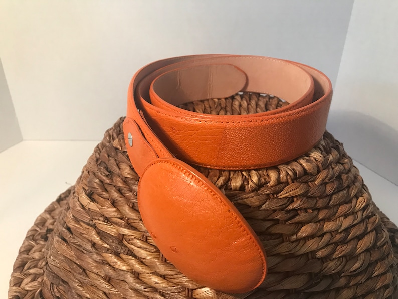 Op de afbeelding: Een feloranje leren riem met een grote, ronde gesp. De riem is opgerold en ligt op een geweven mand.