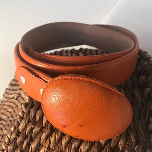 Op de afbeelding: Een feloranje leren riem met een grote ovale gesp. De riem is gevouwen en ligt op een geweven rieten oppervlak.