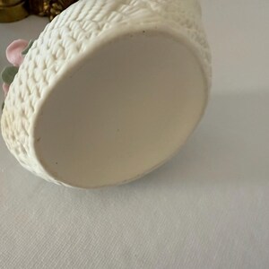 May include: Off-white ceramic swan-shaped container with a textured exterior. The container has a basket weave pattern and is decorated with pink and green floral accents. The bottom of the container is smooth and unadorned.